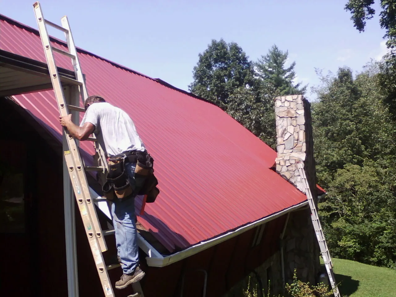 Roofer on a ladder beside a red metal roof and stone chimney — Soffit & Fascia Repair by Roofing Brothers in Willis